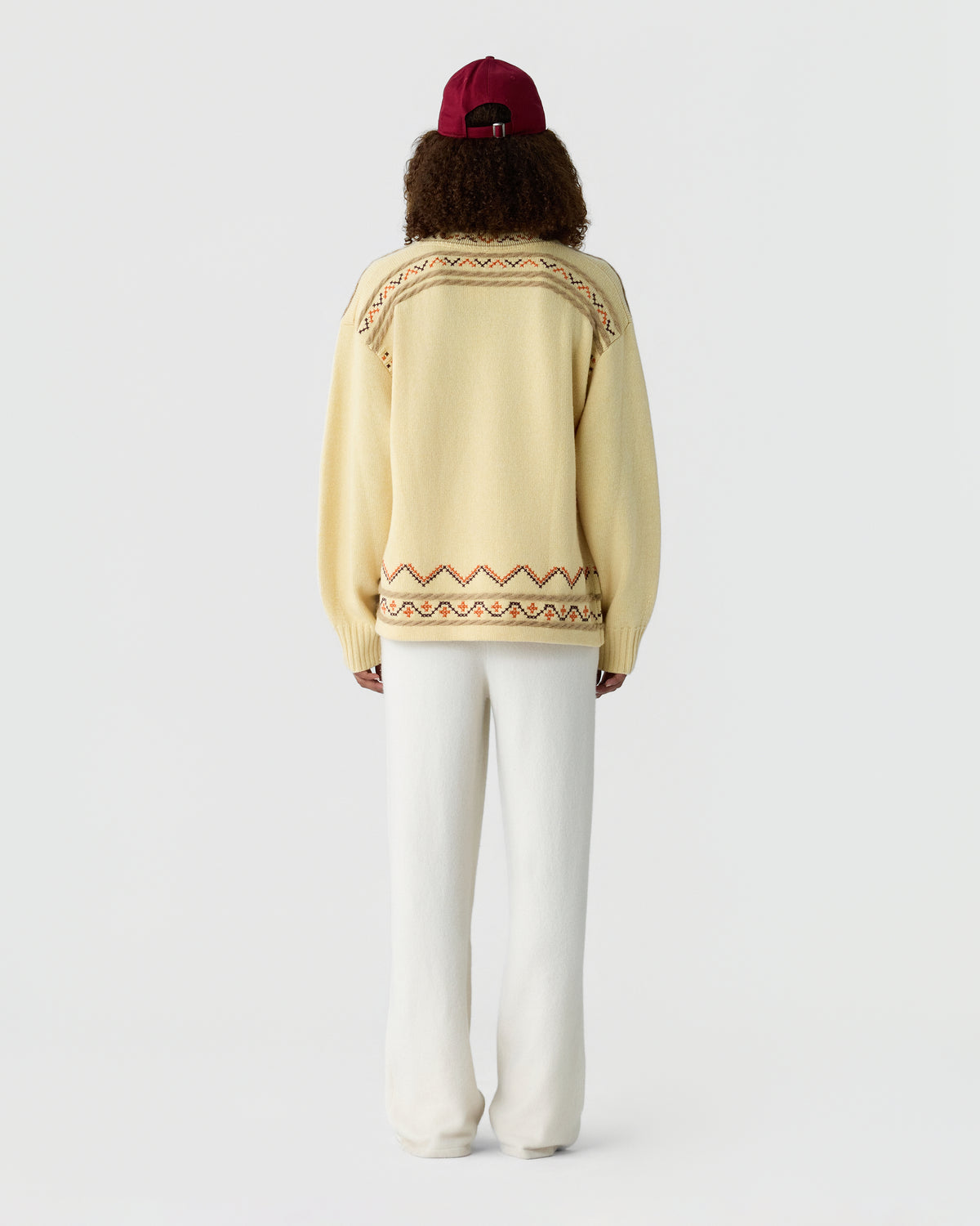 Back view of a person wearing a cream sweater with colorful patterned trim and white trousers with a burgundy cap against a light gray background