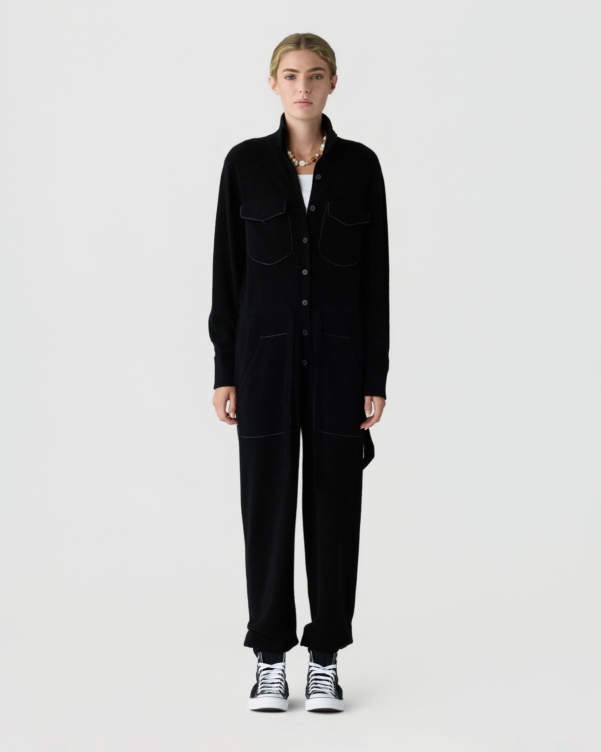 Person standing against a light gray background, wearing an all black long sleeve jumpsuit with chest pockets and white and black sneakers.
