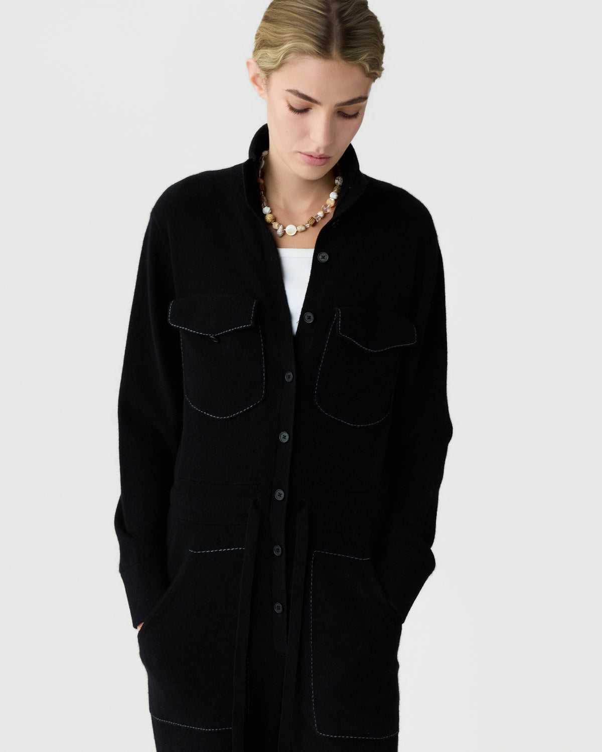 Person wearing a black button front jacket with four patch pockets over a white shirt and a beaded necklace against a light background