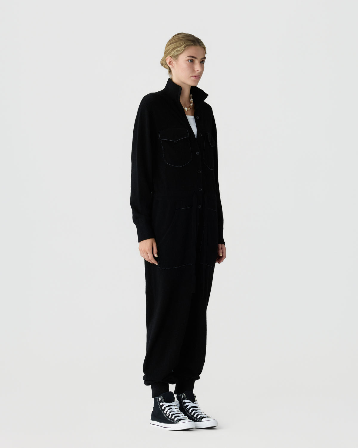 Full length view of a woman wearing a loose black collared jacket and matching pants, paired with black and white high top sneakers.