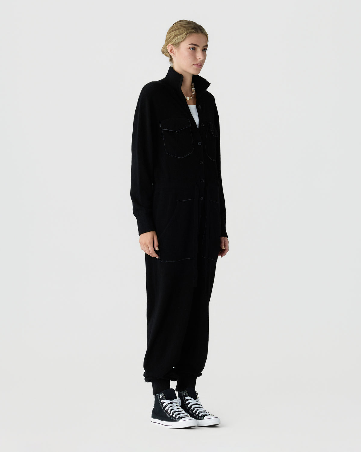 Full length studio portrait of a woman wearing a black utility style jumpsuit with a high collar and black and white high top sneakersաժողով
