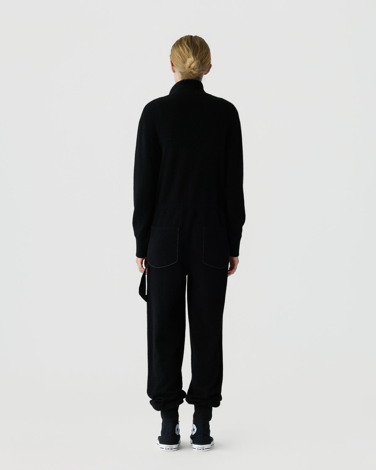 Back view of a model wearing a black high neck sweater and matching relaxed trousers with white and black sneakers against a light gray studio background