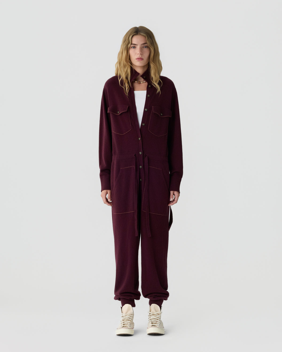 Woman with long wavy hair wearing a burgundy button front jumpsuit with chest pockets and white sneakers against a light gray background