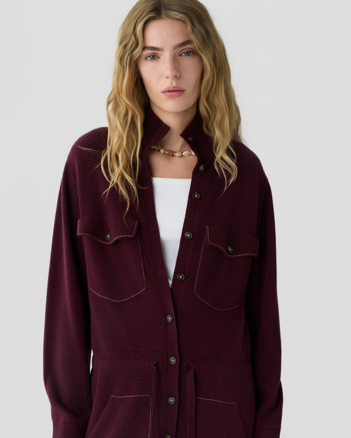 Woman with long wavy blonde hair wearing a burgundy button front jacket with flap pockets over a white top against a light gray background