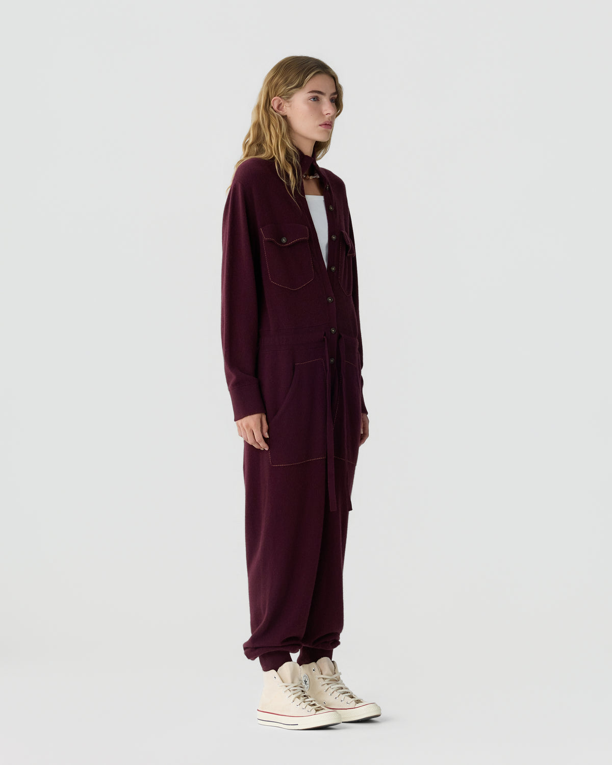 Woman wearing a burgundy utility jumpsuit over a white shirt with cream sneakers against a light gray background