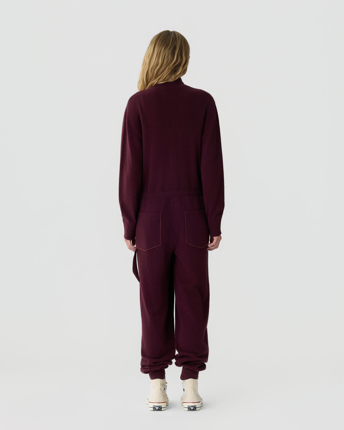 Person viewed from behind wearing a burgundy long sleeve jumpsuit with gathered cuffs and white shoes against a light gray background