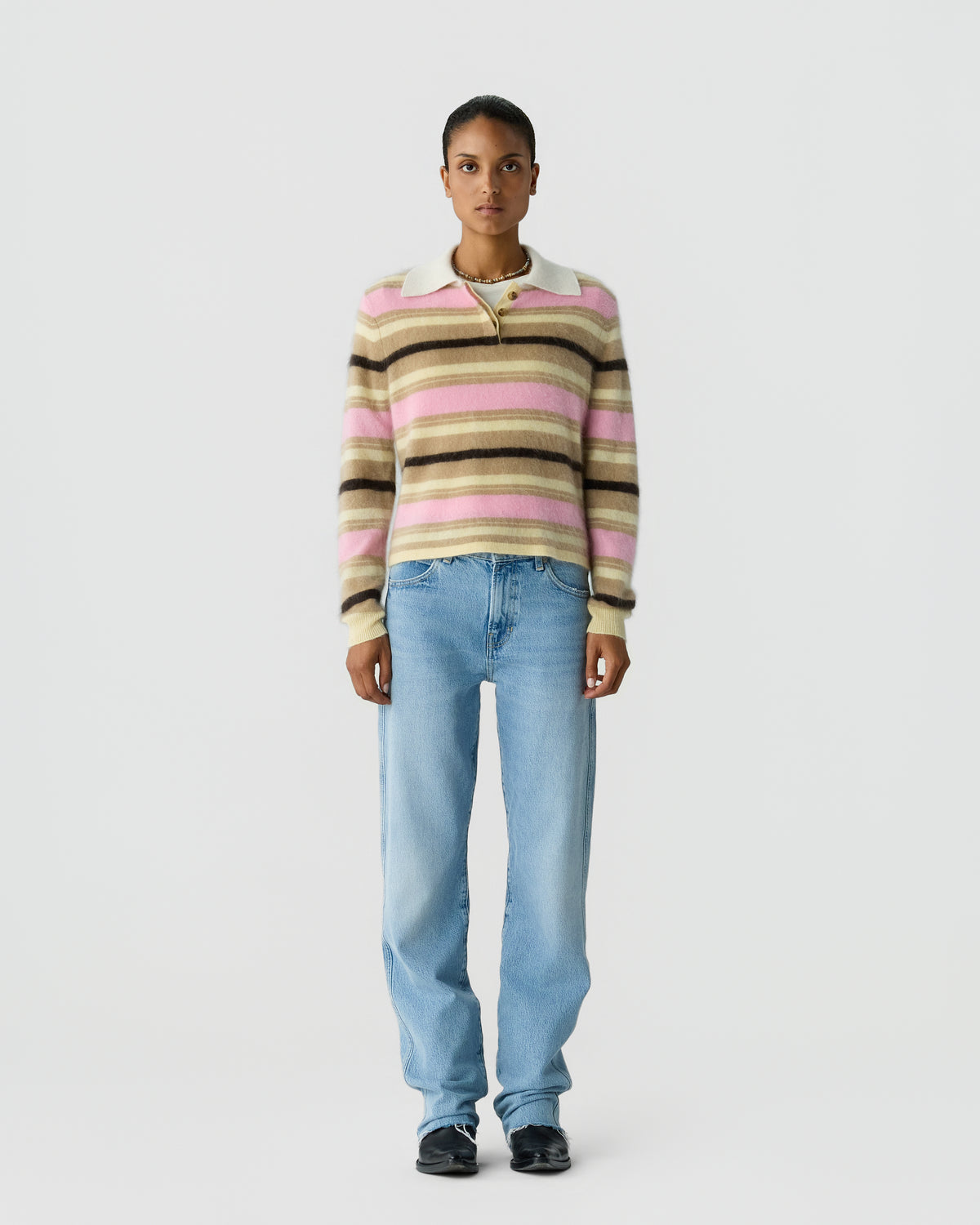 Person wearing a striped pink, beige, and brown collared sweater with light blue jeans and black shoes against a plain light gray background
