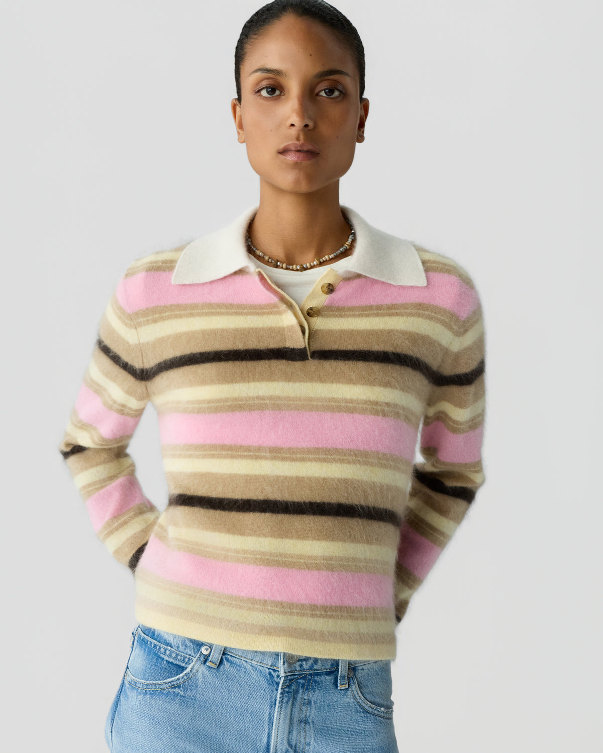 Model wearing a pink, cream, brown, and black striped collared sweater with blue jeans against a light gray background