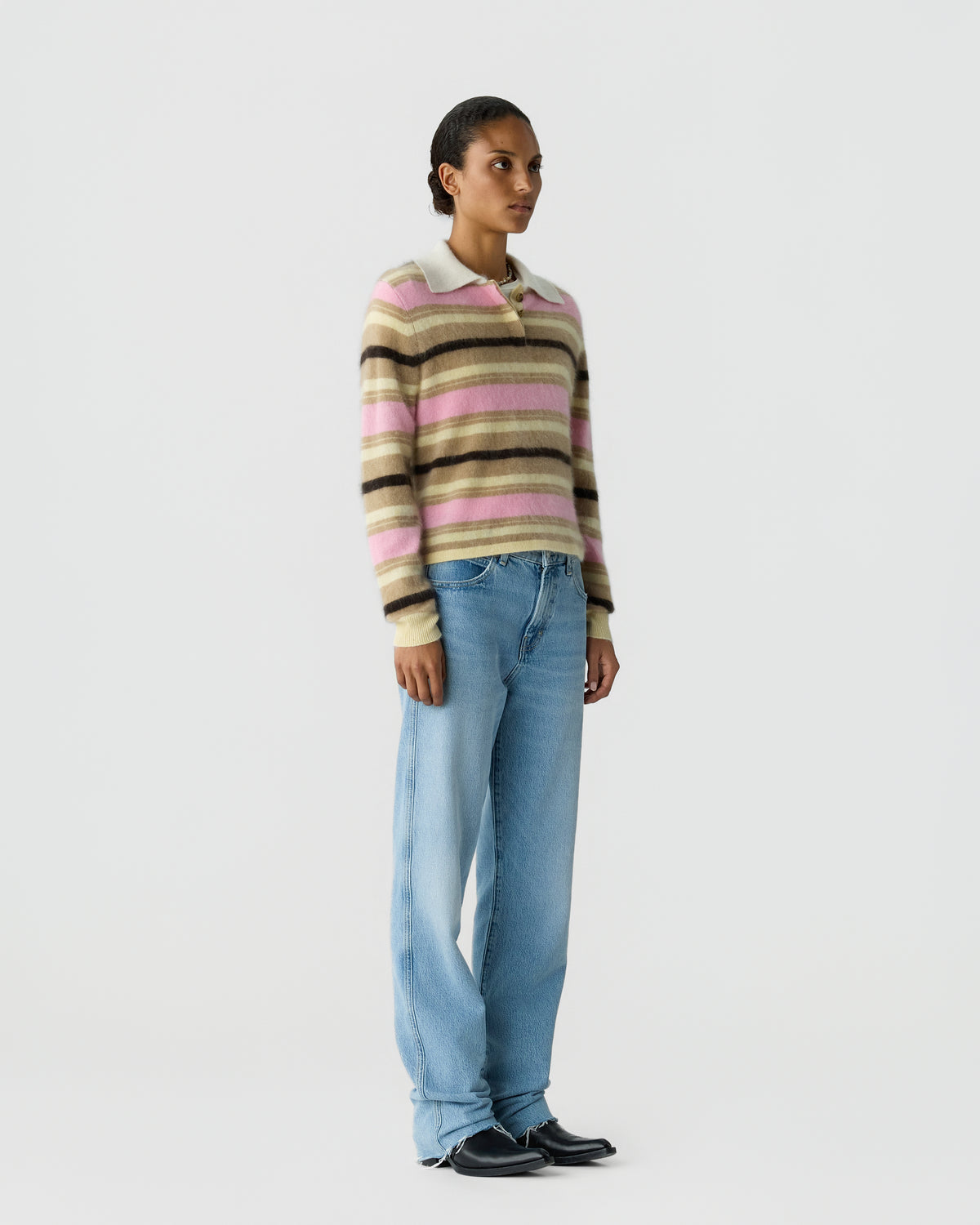 Model wearing a striped cashmere sweater in pink beige and black with blue jeans and black shoes against a light gray studio background