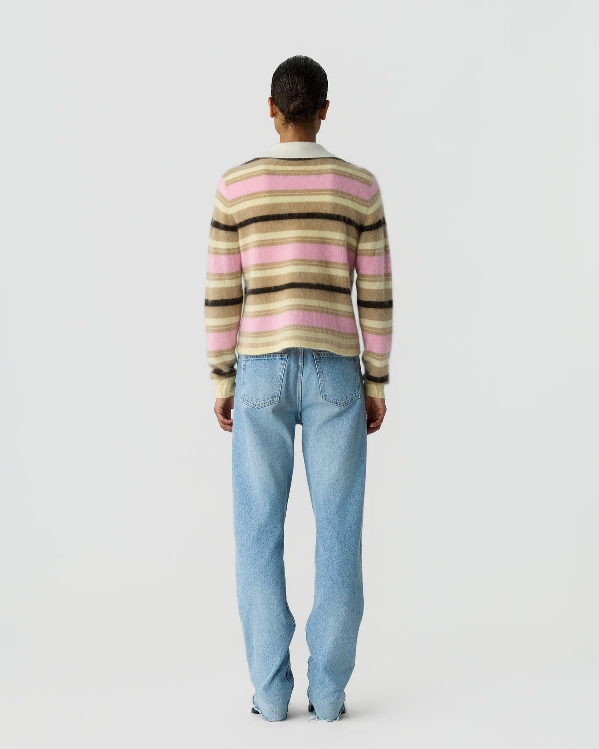 Rear view of a person wearing a striped pink cream and brown knit sweater with light blue jeans against a plain light gray background