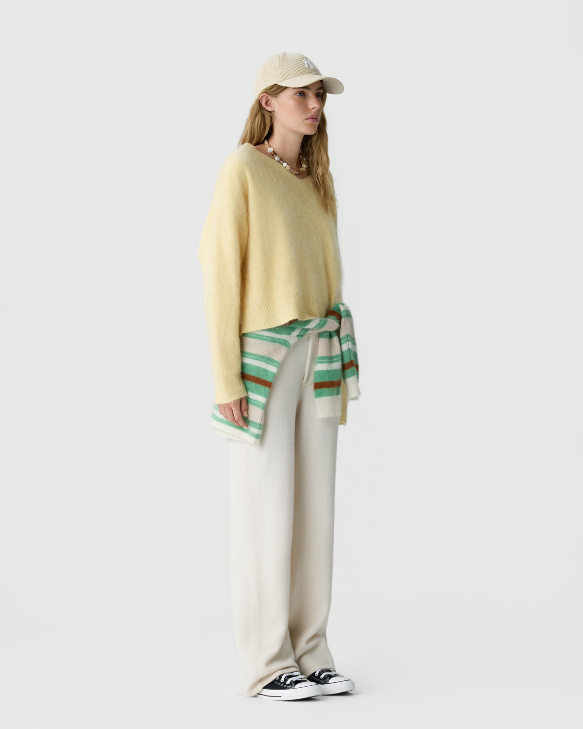 Woman wearing a light yellow cashmere sweater, beige wide-leg pants, a beige baseball cap, black and white sneakers, and a striped green and beige sweater tied around her waist against a plain background.