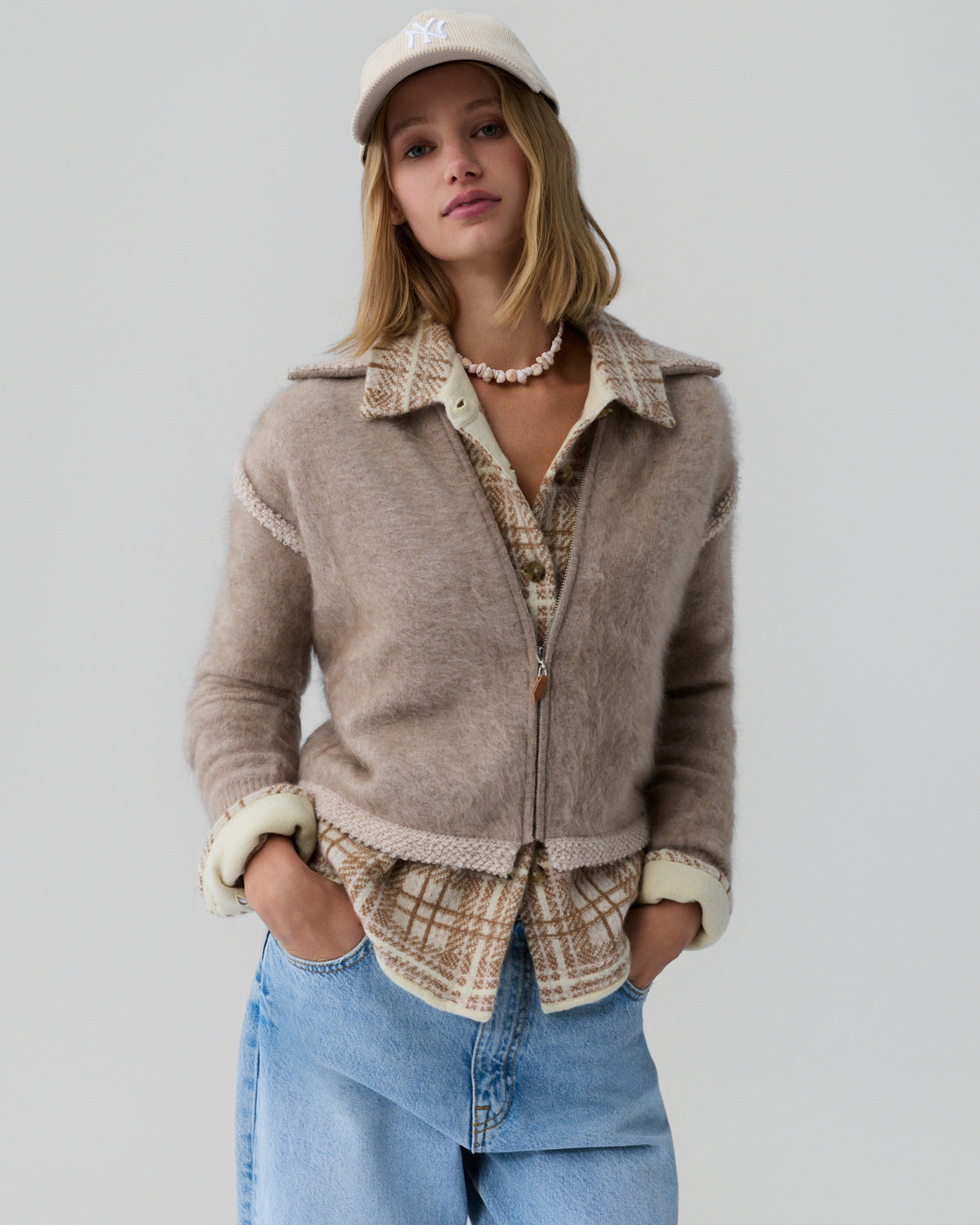 Young woman wearing a soft taupe fuzzy zip-up jacket layered over a beige plaid button-up shirt, light blue denim jeans, a cream baseball cap, and a shell necklace against a plain light gray background.