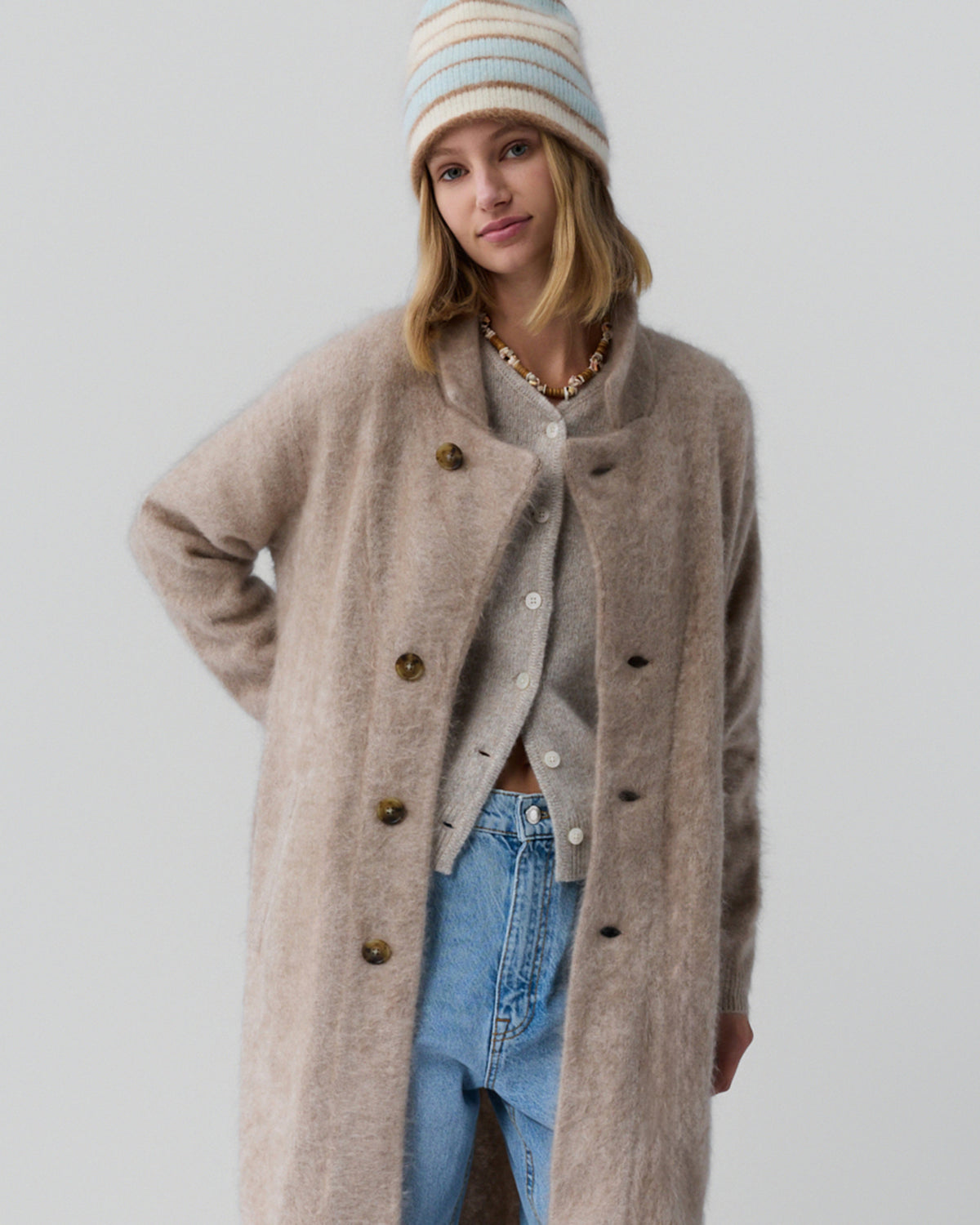 Young woman wearing a soft taupe long coat with large buttons layered over a light gray cardigan and light blue jeans, accessorized with a striped knit beanie and a shell necklace against a plain background.
