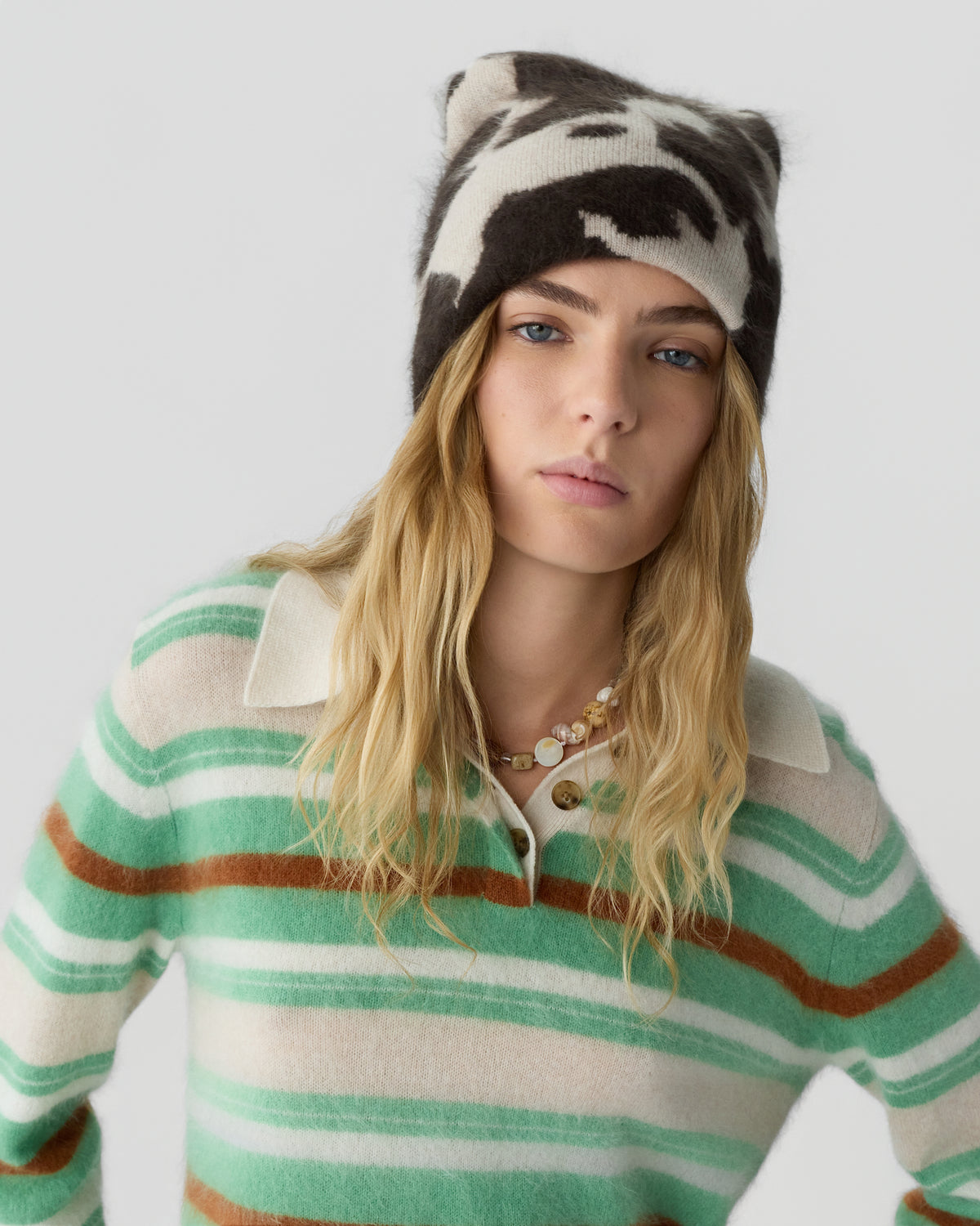 Young woman with long blonde hair wearing a black and white patterned beanie and a green, brown, and cream striped collared sweater against a plain light gray background.