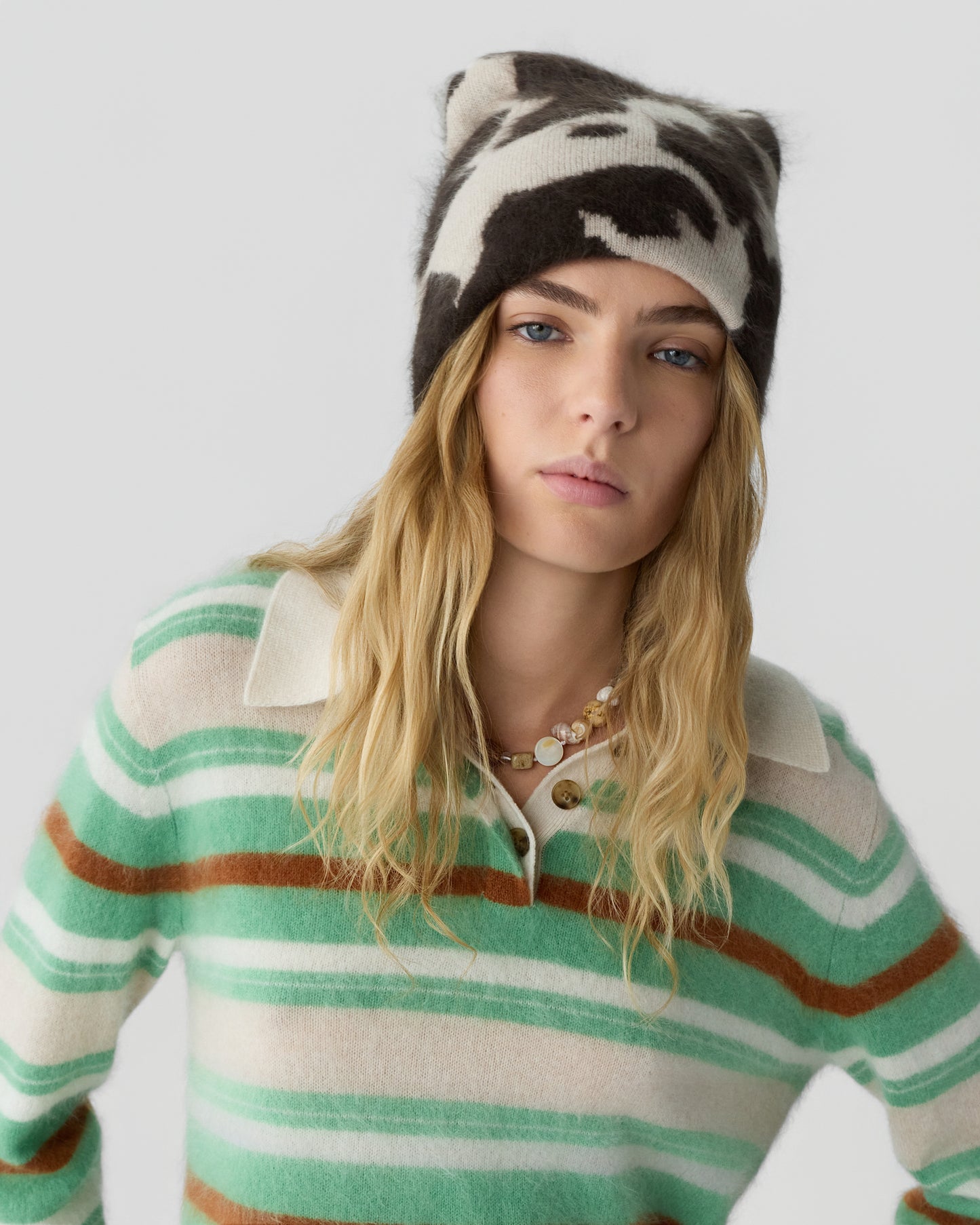 Young woman with long blonde hair wearing a black and white patterned beanie and a green, brown, and cream striped collared sweater against a plain light gray background.