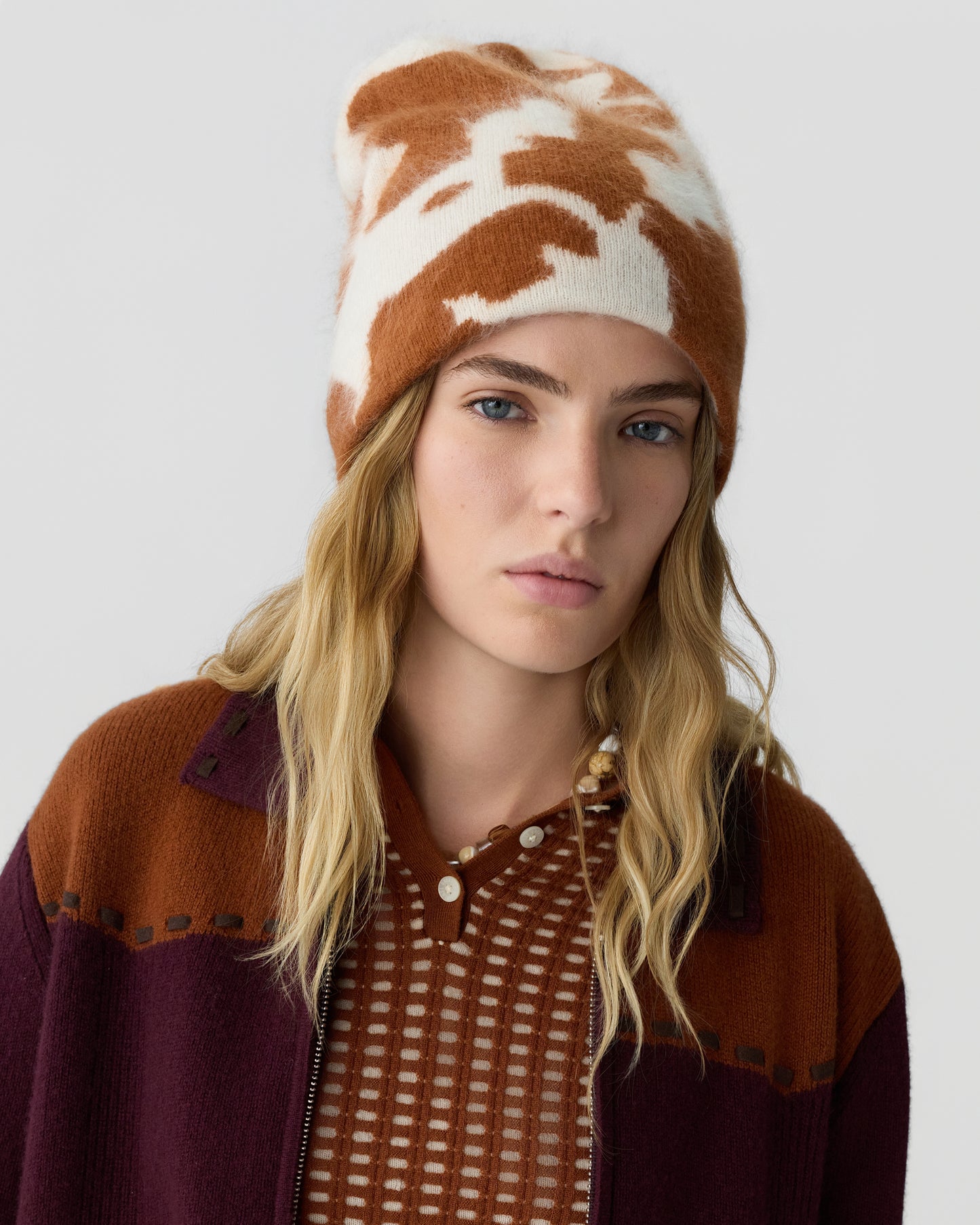 Young woman with long blonde hair wearing a brown and white patterned beanie and a multicolored jacket over a textured blouse against a gray background