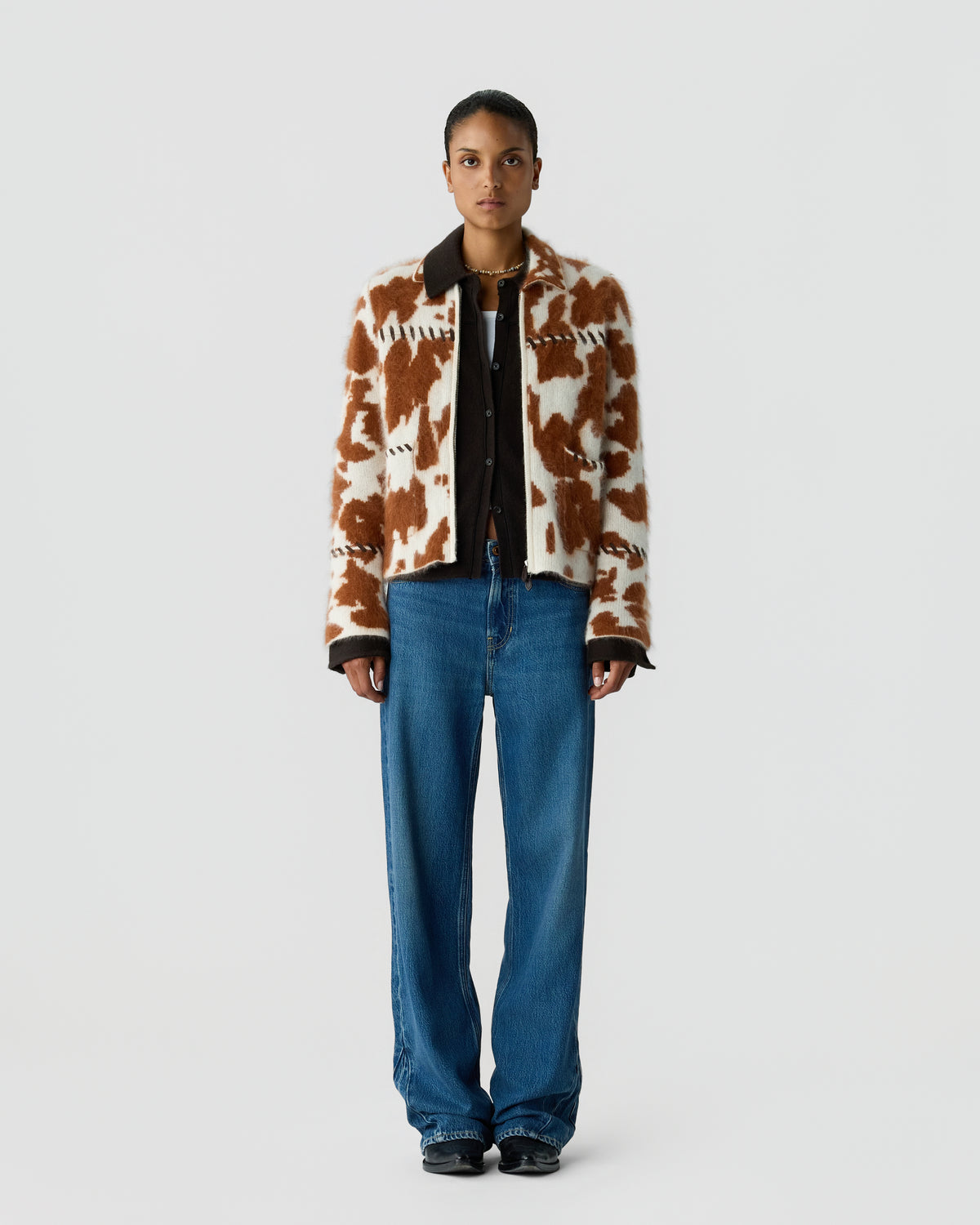 Person standing against a light gray background wearing a brown and cream patterned cardigan over a black shirt with blue jeans and black shoes