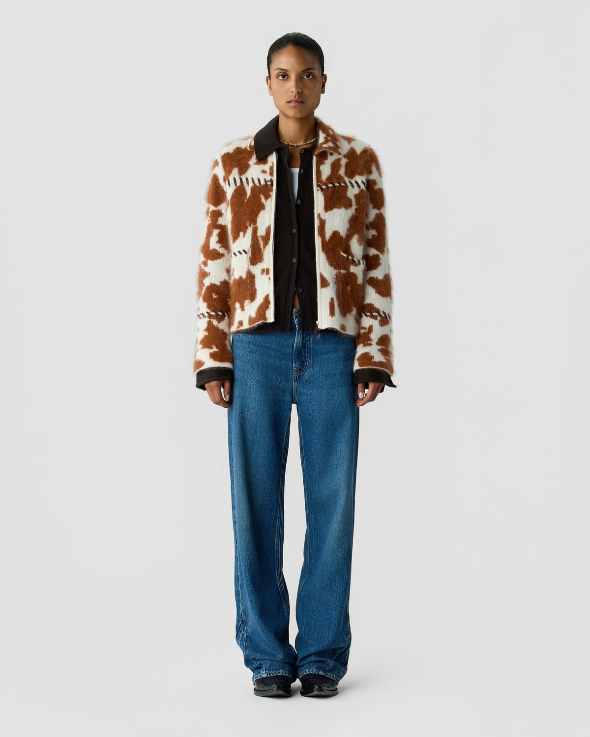 Model wearing a brown and cream patterned cardigan over a black top, with blue jeans and black shoes against a plain light gray background