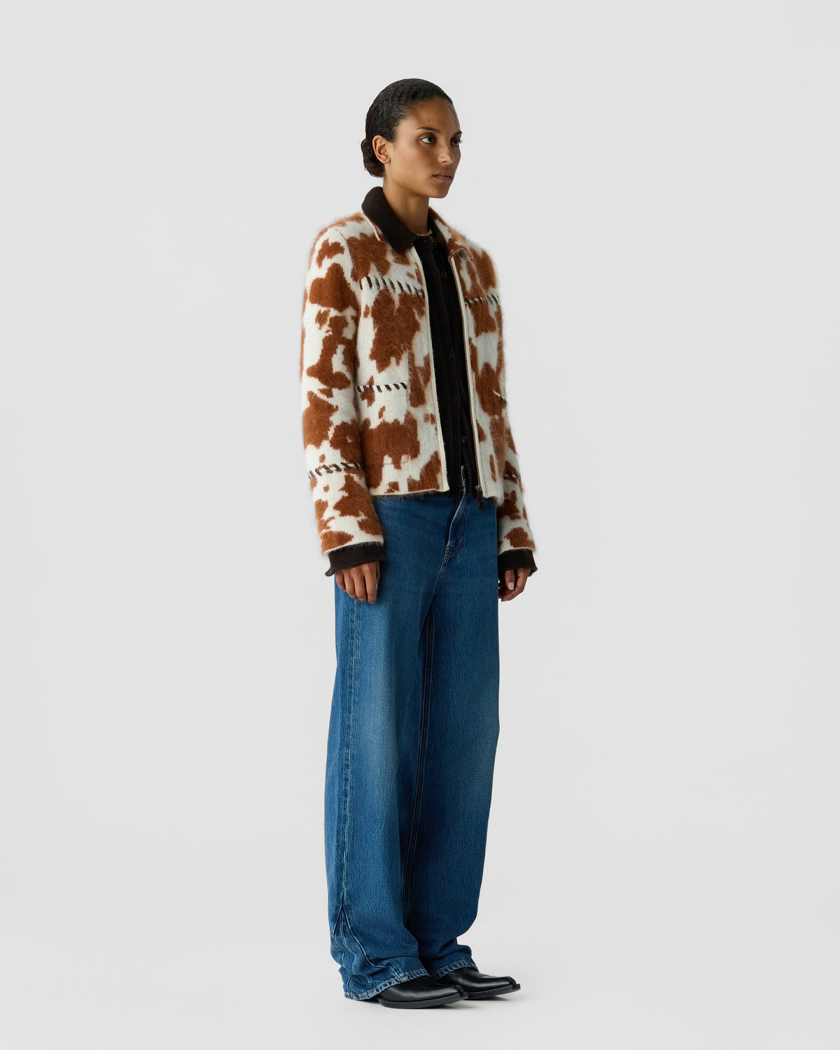 Model wearing a brown and cream cow print cardigan over a dark shirt, wide leg blue jeans, and black shoes against a light gray studio background