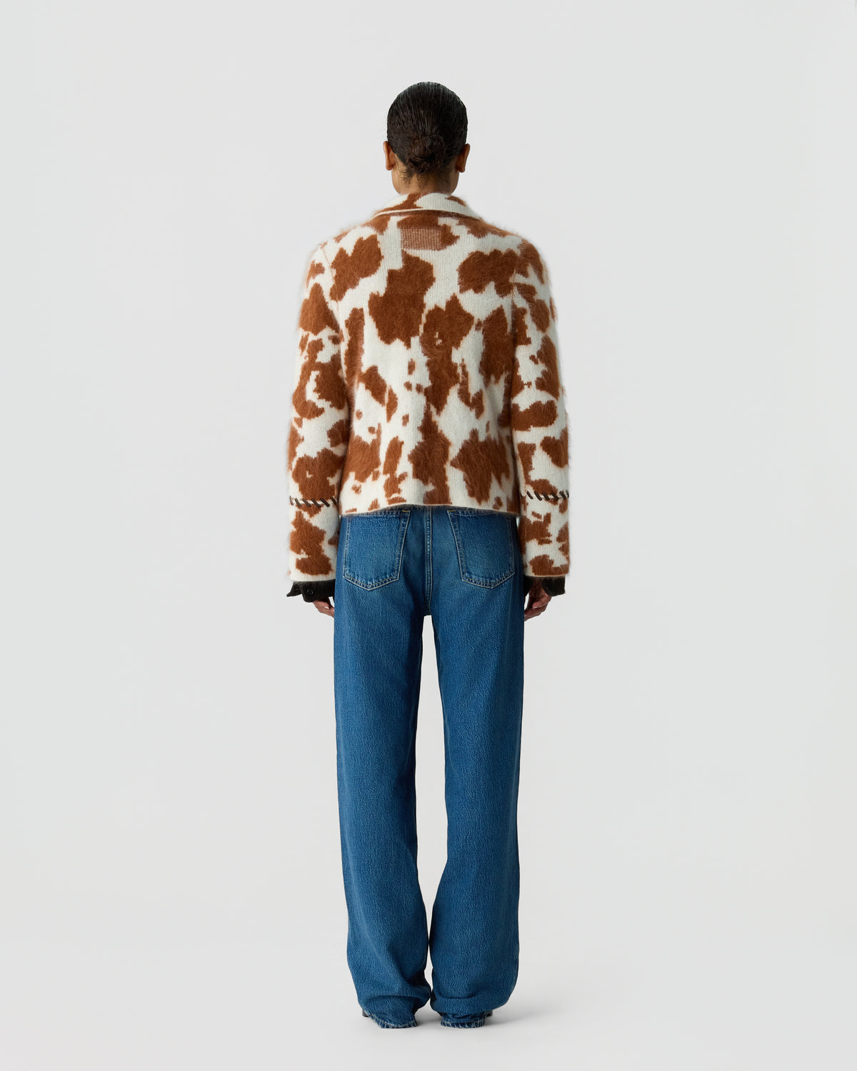 Person viewed from behind wearing a brown and cream patterned sweater with blue jeans against a plain light background