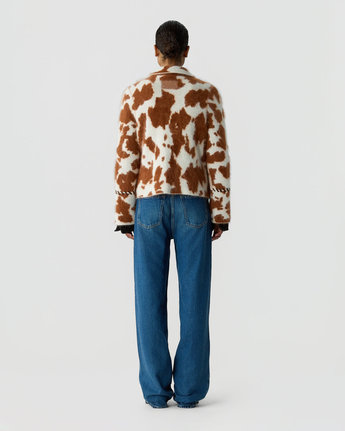 Back view of a person wearing a cream and brown patterned sweater with blue jeans against a plain light gray background
