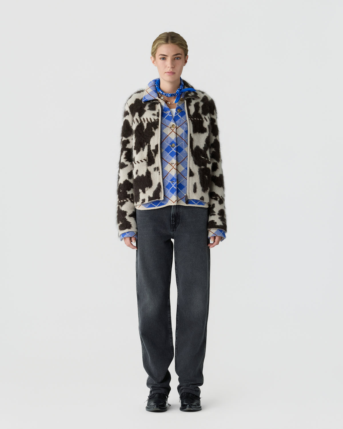 Full length portrait of a person wearing a black and white patterned cardigan over a blue plaid shirt with dark jeans and black shoes against a light gray background