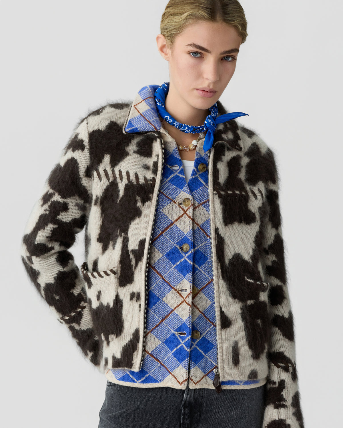 Fashion model wearing a black and white patterned cardigan over a blue argyle shirt with a matching neck scarf and layered necklaces against a gray background