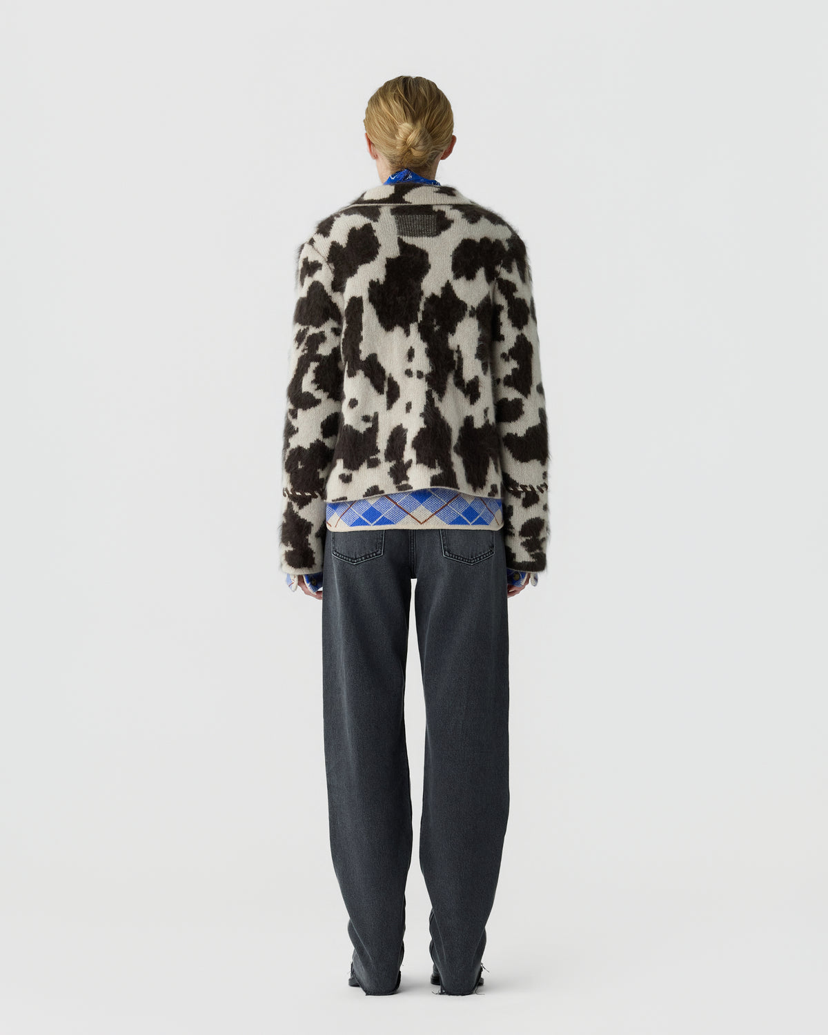 Person viewed from behind wearing a black and white cow print sweater with dark relaxed trousers against a light gray studio background
