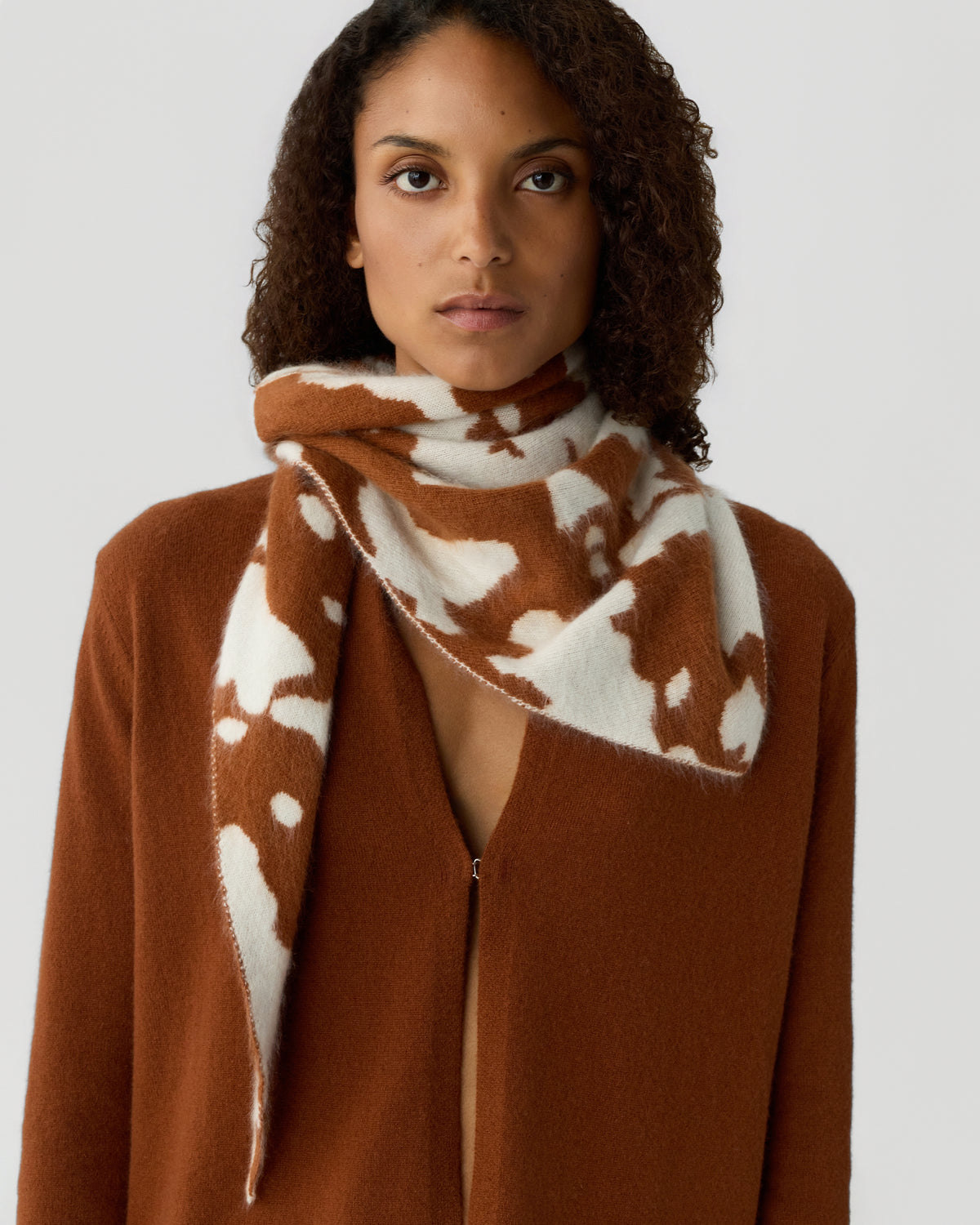 Woman wearing a rust-colored open-front cardigan and a matching scarf with a white and brown abstract pattern wrapped around her neck against a plain background.