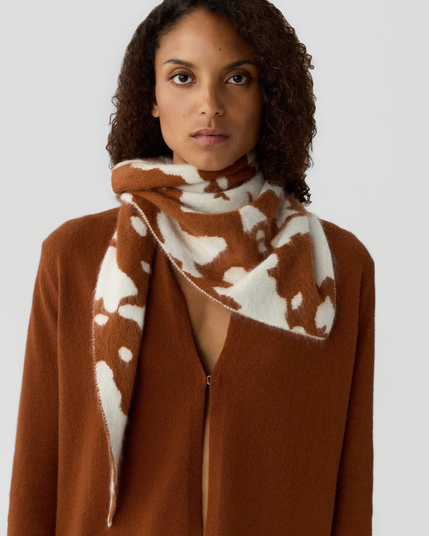Woman wearing a rust-colored open-front cardigan and a matching scarf with a white and brown abstract pattern wrapped around her neck against a plain background.
