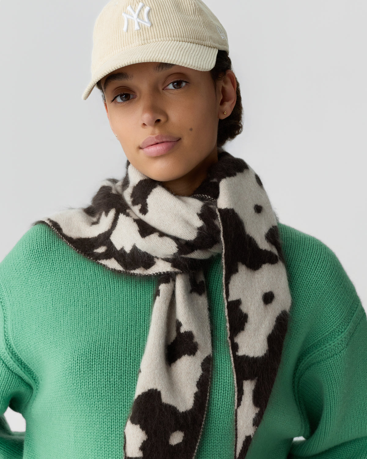 Woman wearing a beige cap, green knit sweater, and a black and white patterned scarf wrapped around her neck against a plain background.