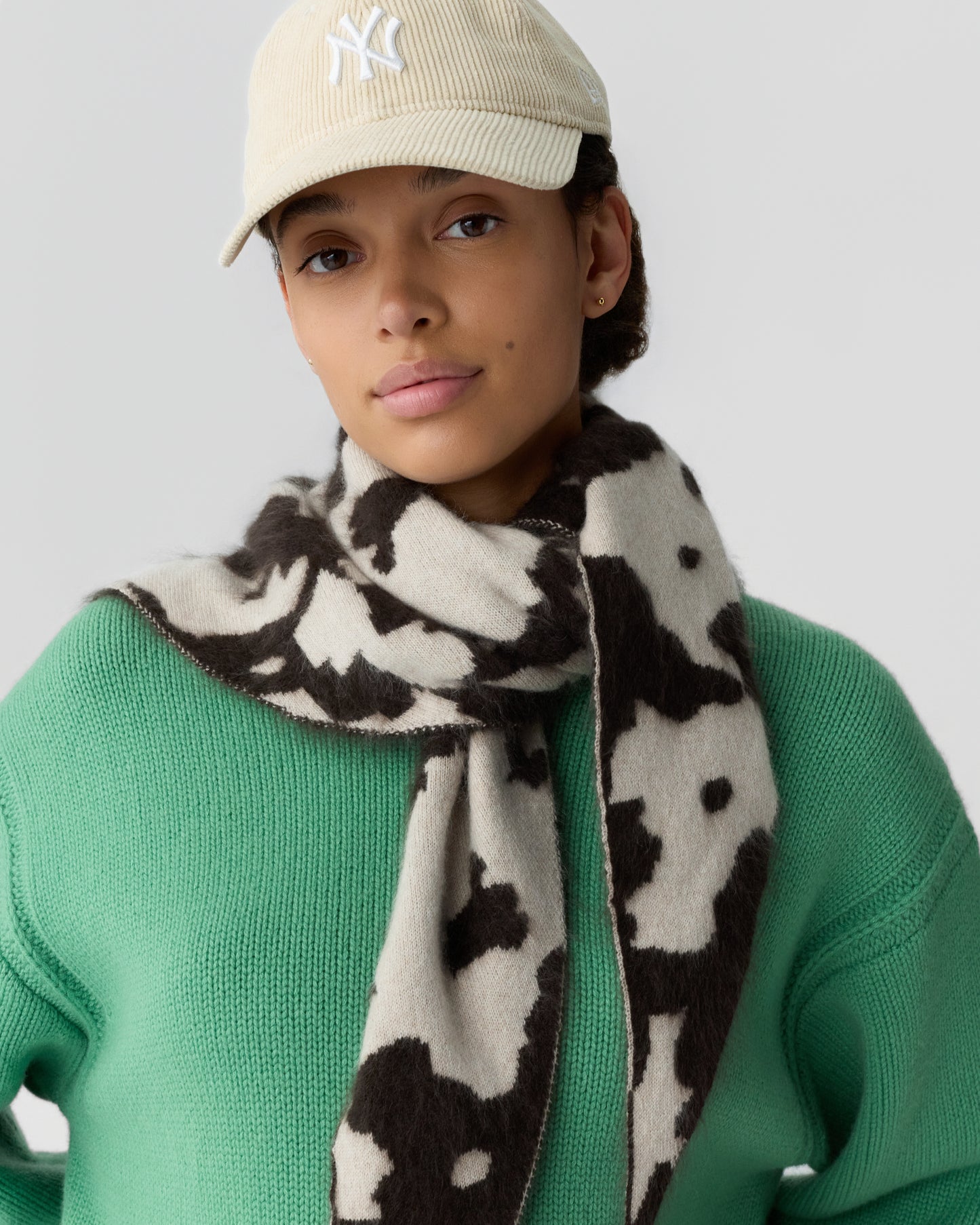 Woman wearing a beige cap, green knit sweater, and a black and white patterned scarf wrapped around her neck against a plain background.