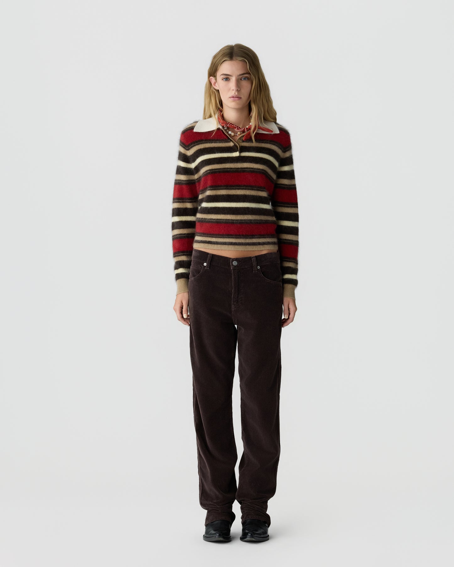 Young woman standing against a plain white background wearing a striped sweater with red, beige, and black horizontal lines, dark brown pants, and black shoes.