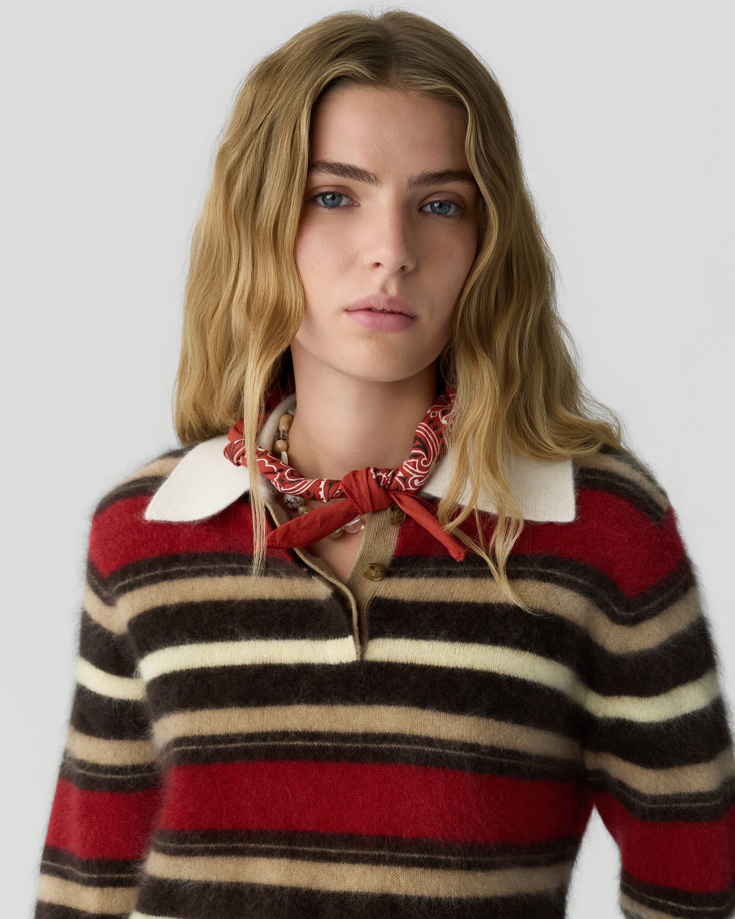 Young woman with long wavy blonde hair wearing a striped fuzzy sweater in red, brown, beige, and white with a white collar, and a red patterned neck scarf.
