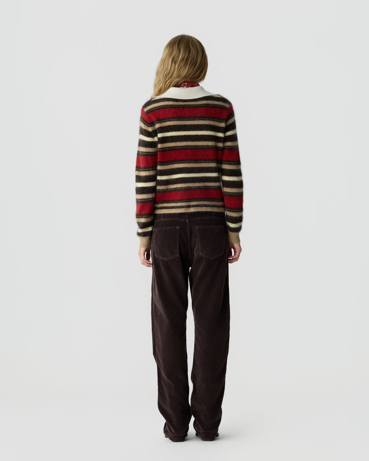 Woman seen from behind wearing a long-sleeve striped sweater with red, black, and beige colors paired with loose dark brown pants against a plain gray background.