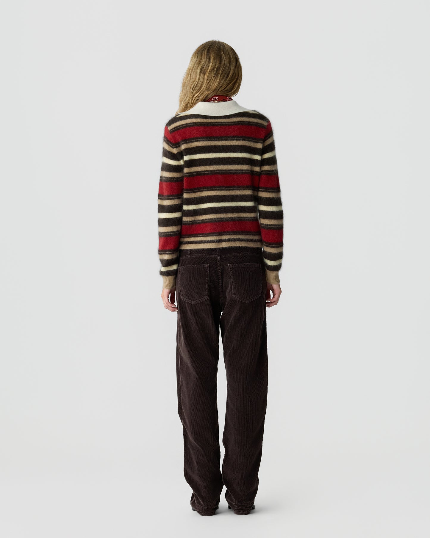 Woman seen from behind wearing a long-sleeve striped sweater with red, black, and beige colors paired with loose dark brown pants against a plain gray background.