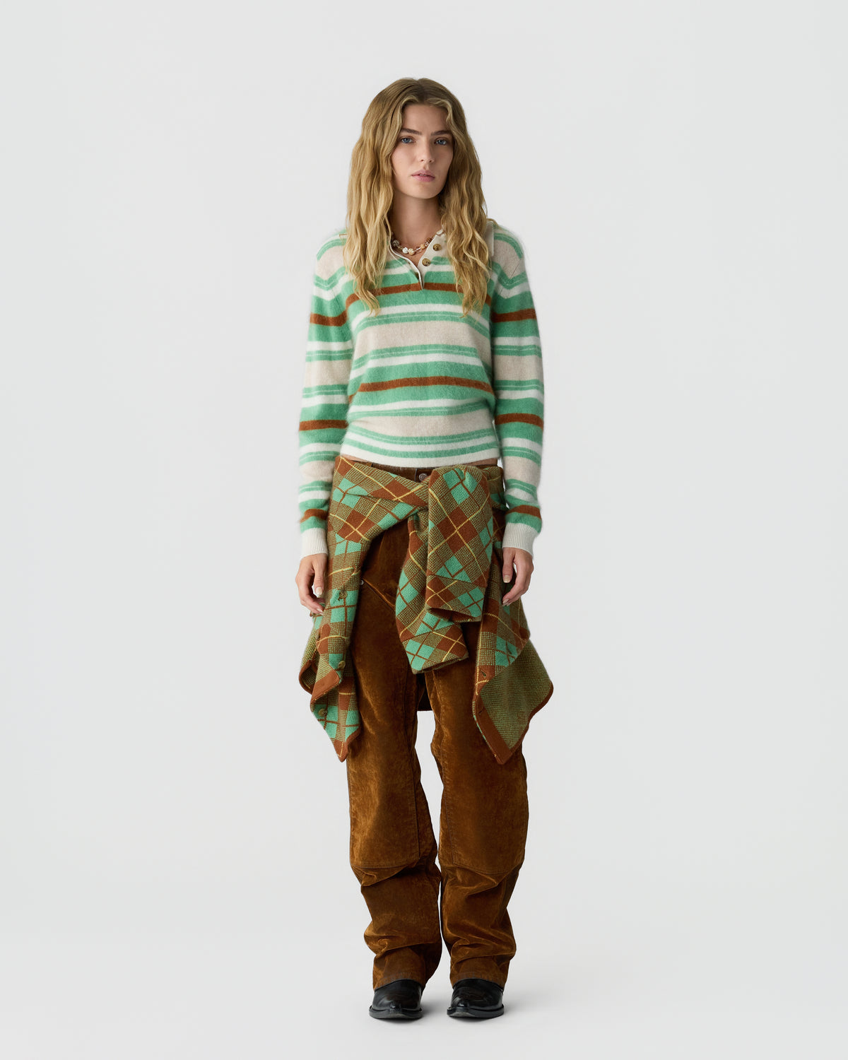 Young woman with wavy blonde hair wearing a green, white, and brown striped cashmere sweater, brown pants, and a green and brown plaid sweater tied around her waist.