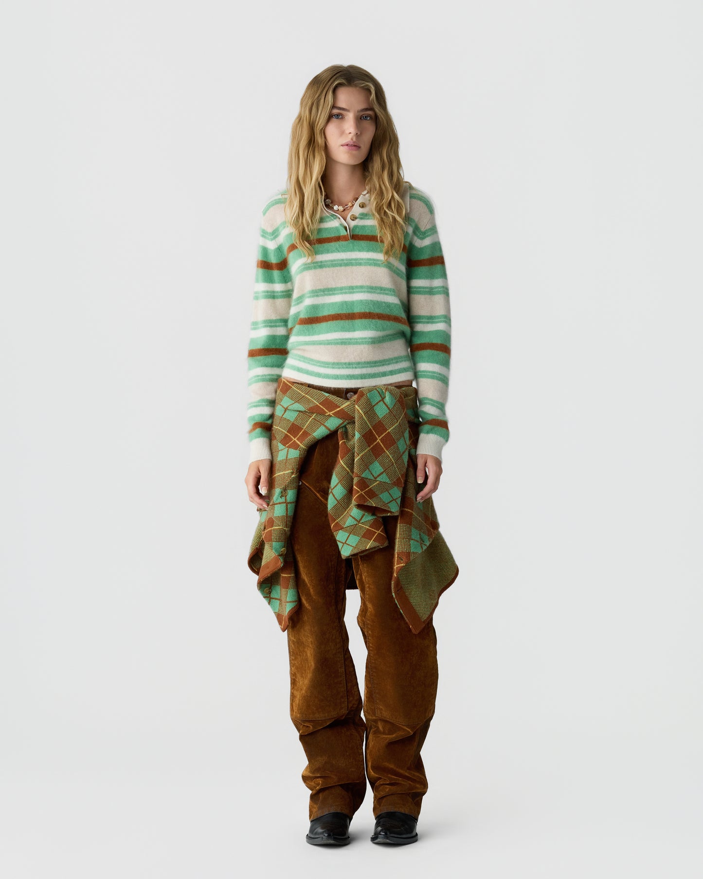 Young woman with wavy blonde hair wearing a green, white, and brown striped cashmere sweater, brown pants, and a green and brown plaid sweater tied around her waist.