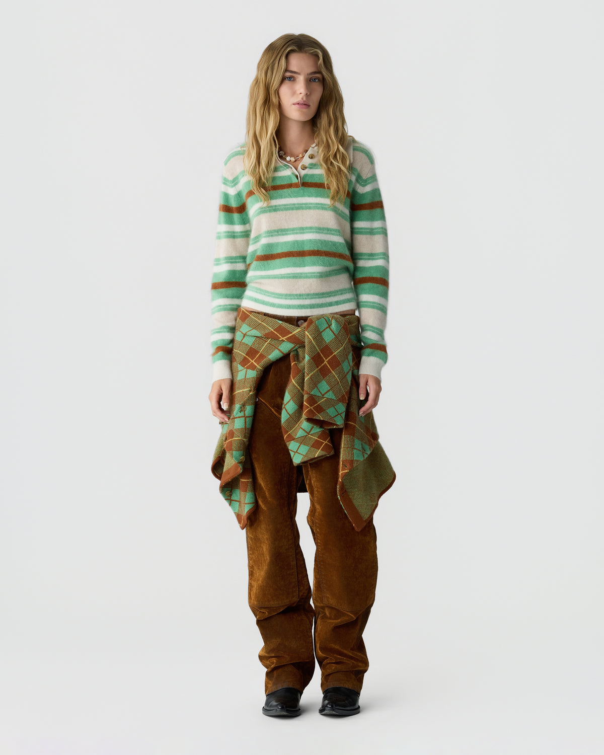 Model wearing a green and cream striped knit sweater, patterned brown draped garment, wide brown trousers, and black shoes against a light background
