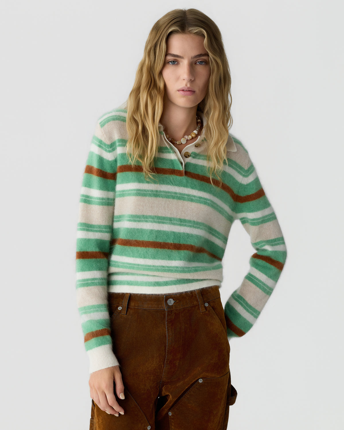 Young woman with long blonde hair wearing a striped green, white, and brown sweater with brown pants against a plain light background.