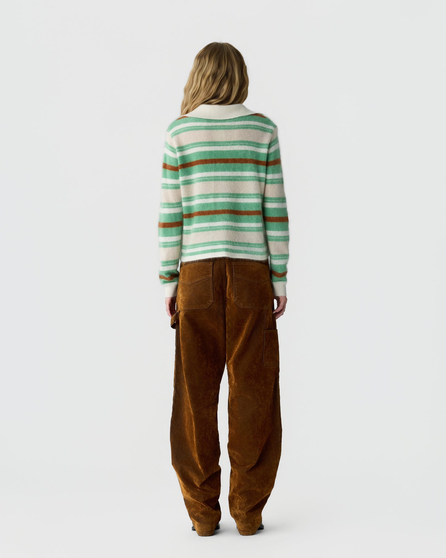 Woman seen from the back wearing a striped sweater with green, white, and brown horizontal lines and loose-fitting brown corduroy pants against a plain background.