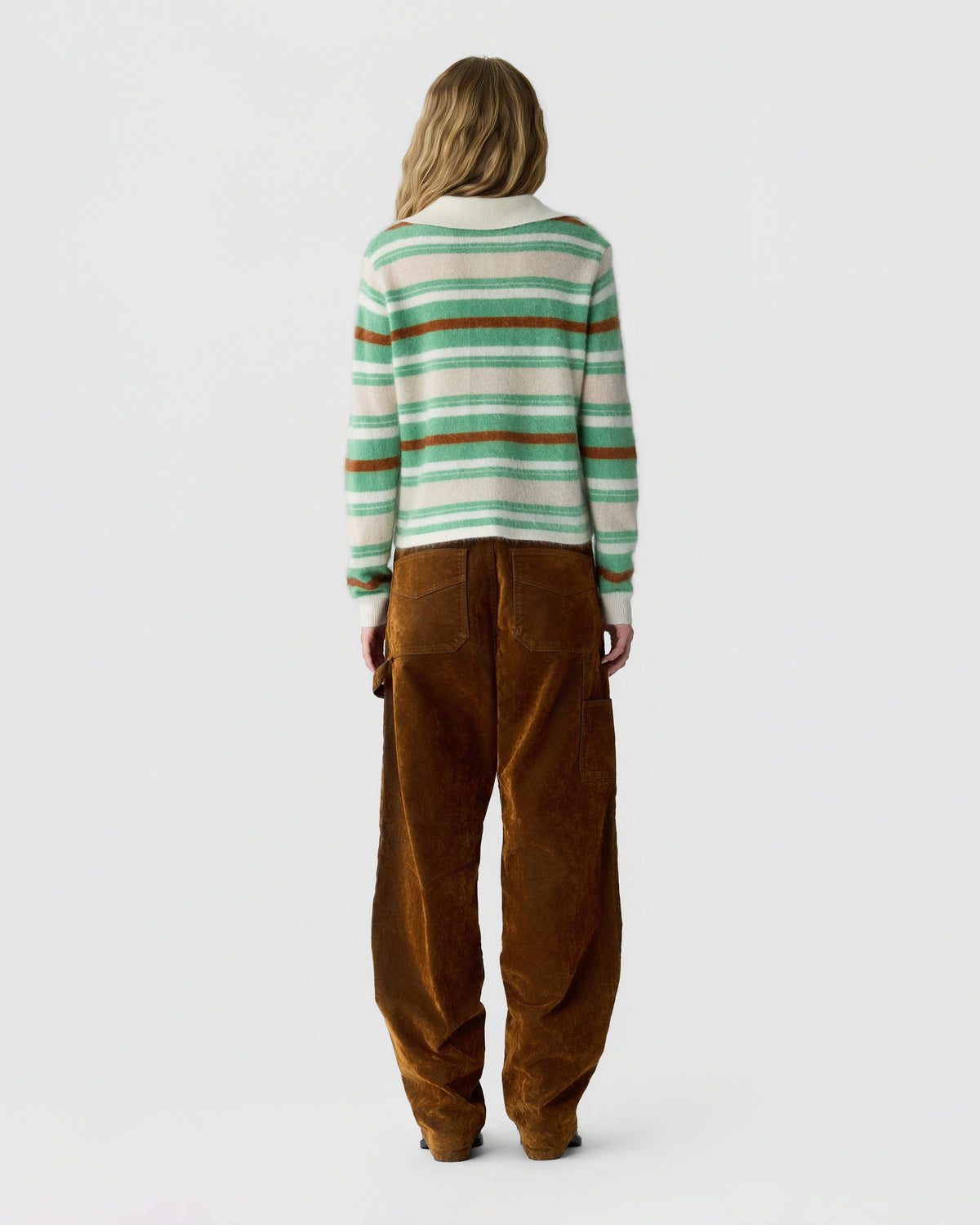 Rear view of a person wearing a green and cream striped sweater with a red stripe, loose brown trousers, and dark shoes against a light background