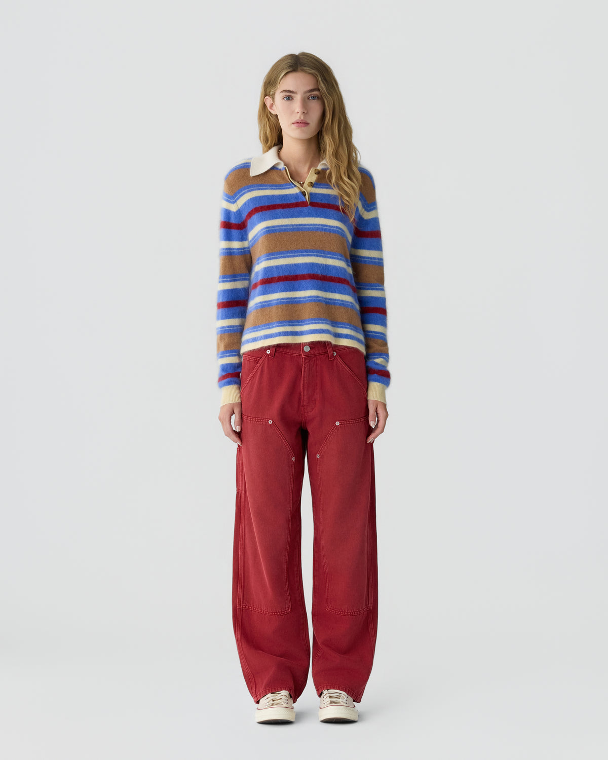 Model wearing a blue red and cream striped collared sweater with red wide leg trousers and light sneakers against a plain pale gray background