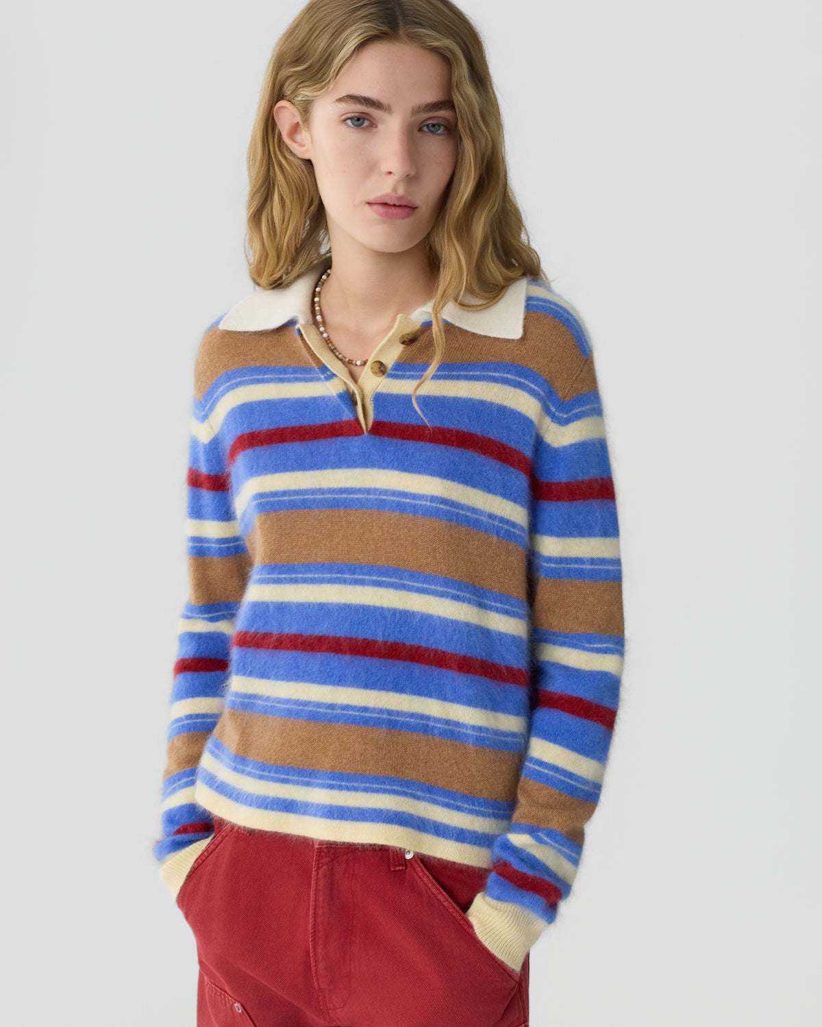 Young woman wearing a blue tan red and cream striped sweater with a white collar and red trousers against a light gray background