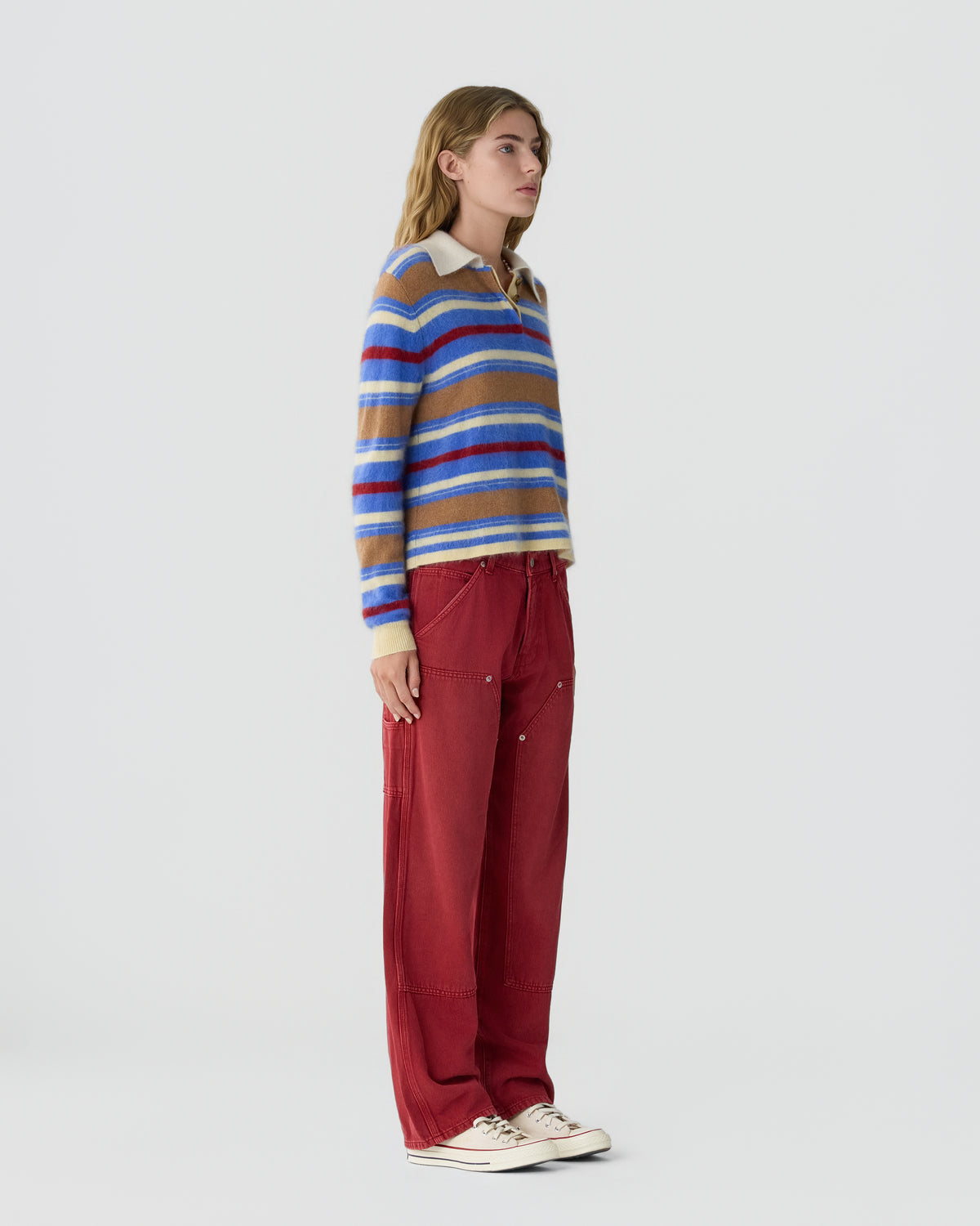 Model wearing a striped blue red and tan sweater with loose red trousers and white sneakers against a light gray studio background