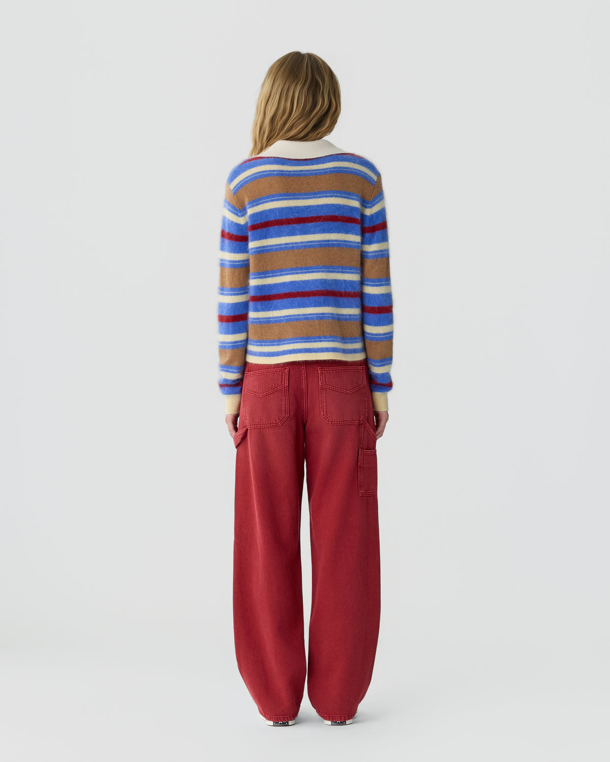 Person viewed from behind wearing a blue, red, and tan striped sweater with loose red pants against a plain light gray background