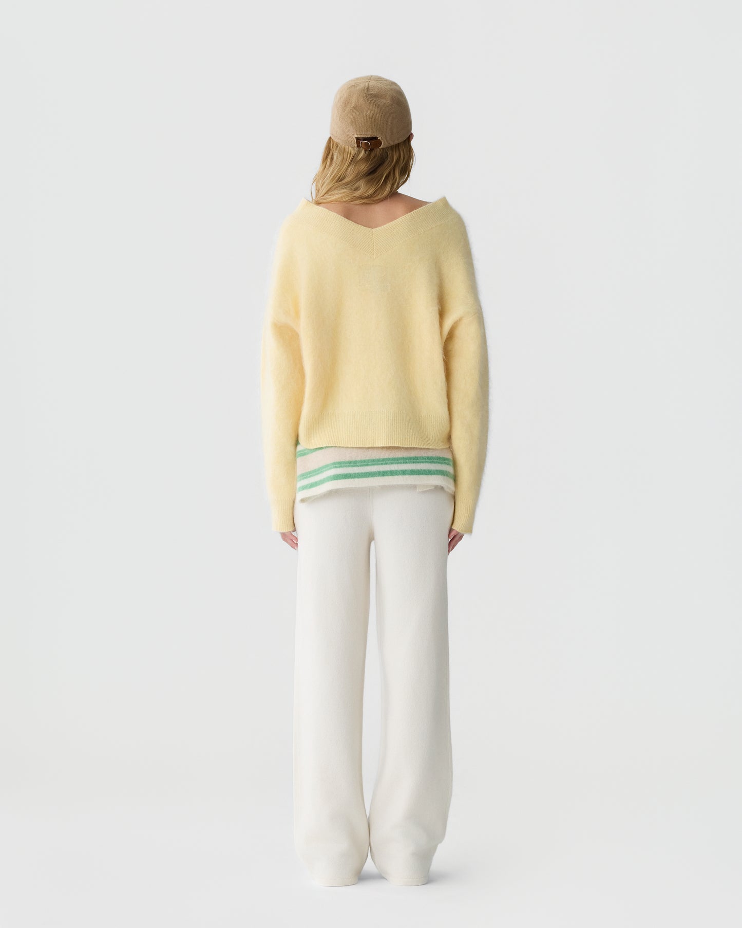 Person seen from behind wearing a soft yellow sweater with a V-shaped back neckline, white pants, a striped shirt peeking out, and a beige cap against a plain background.