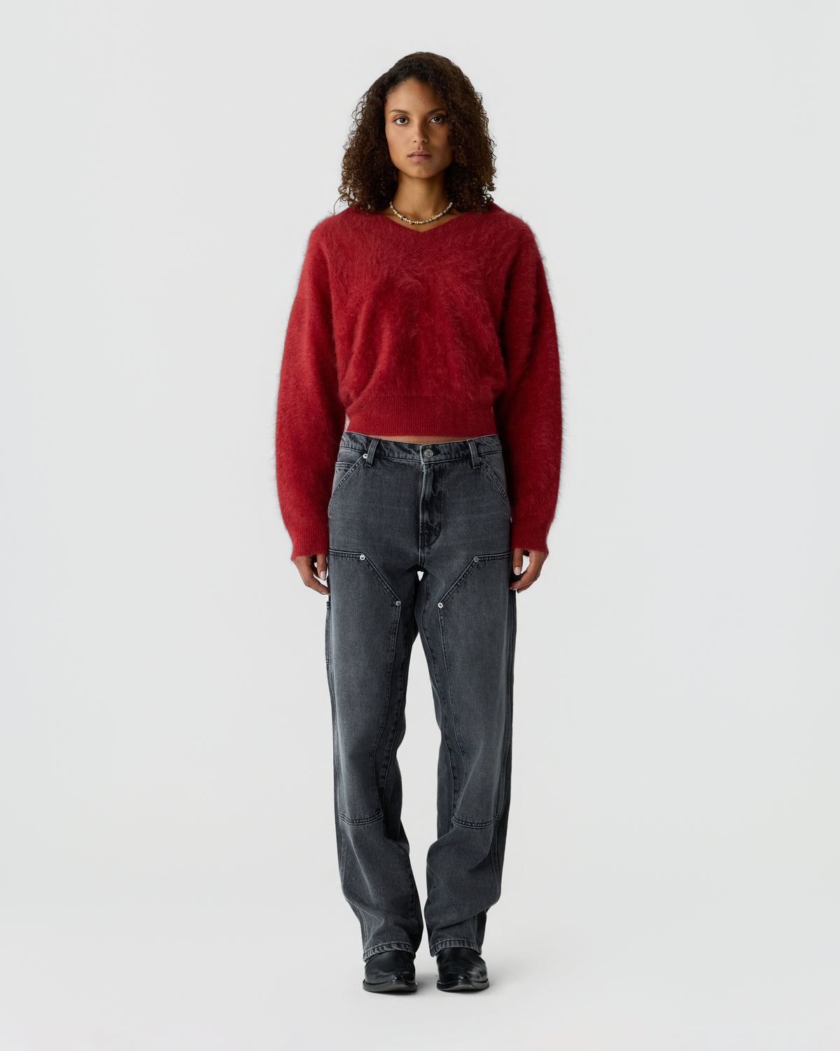 Model wearing a red cashmere sweater with dark gray jeans and black shoes against a light gray background.
