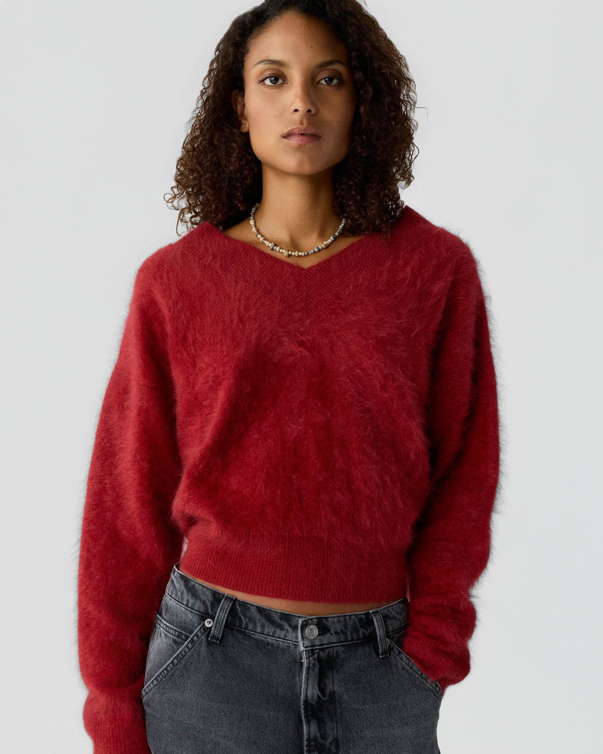 Woman with curly hair wearing a red fuzzy knit sweater, layered necklace, and dark gray jeans against a plain light background