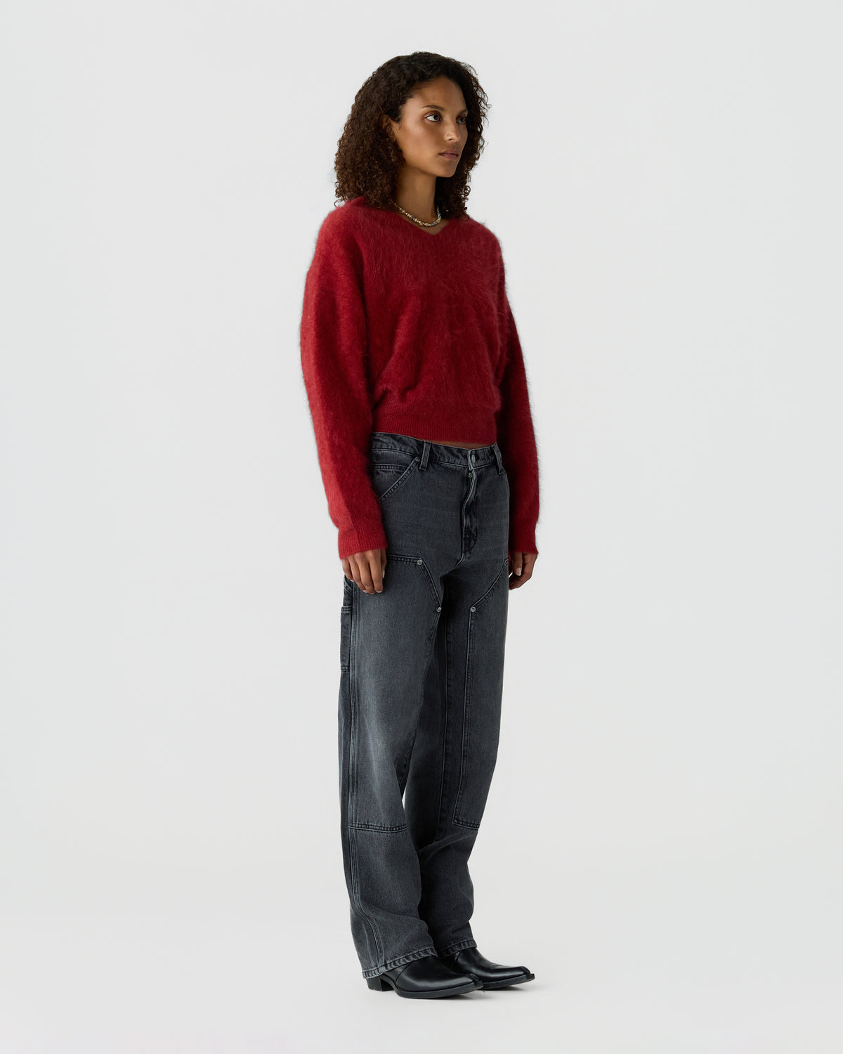 Model wearing a red sweater with dark wide leg jeans and black pointed boots against a light gray studio background
